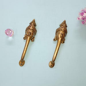 Peut inclure: Deux poignées de porte antiques en laiton avec des sculptures de visages détaillées. Les poignées ont un design long et incurvé et sont posées sur un fond bleu clair. Un petit bouton en verre irisé et une grappe de fleurs roses sont également visibles.