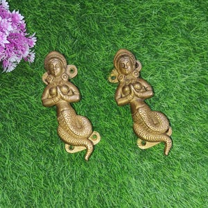 May include: Two gold-toned door handles shaped like mermaids with their hands clasped in prayer. The mermaids are depicted with long flowing hair and are adorned with jewellery.