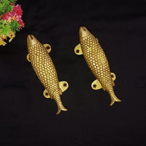 Peut inclure: Deux poignées de porte en forme de poisson de couleur dorée. Les poignées sont détaillées avec des écailles et ont une forme légèrement incurvée.