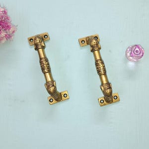 Puede incluir: Dos manijas de puerta de latón antiguo con detalles ornamentados y un pomo de cristal decorativo sobre una superficie azul claro. Las manijas tienen una base rectangular con dos orificios para tornillos y un agarre central texturizado.