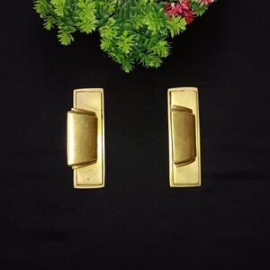 Puede incluir: Dos tiradores de puerta de color dorado con forma rectangular y un diseño curvo en relieve. Los tiradores están sobre un fondo negro.