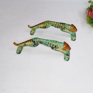 May include: Two metal drawer pulls shaped like leaping tigers. Each handle features a textured, mottled finish in shades of teal, green, and gold, with a smooth, polished gold-tone finish on the tiger's head and front paws for mounting.