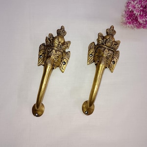 Può includere: Due maniglie per porte in ottone con un design a testa di elefante. Le maniglie sono di colore oro e presentano dettagli intricati, tra cui una corona e motivi decorativi. Le maniglie sono lunghe circa 20 cm.
