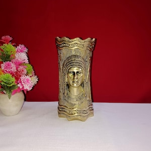 Op de afbeelding: Een gouden metalen vaas met een decoratief ontwerp met een vrouwen gezicht. De vaas heeft een getextureerd oppervlak en een golvende rand.