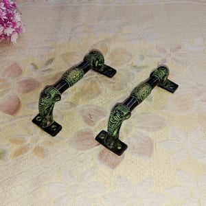 Peut inclure: Deux poignées de porte en métal noir et vert avec des motifs complexes. Les poignées sont en laiton et ont un style traditionnel.