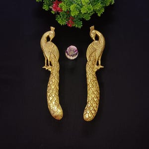 Puede incluir: Dos esculturas de pavo real doradas con patrones de plumas detallados. Los pavos reales están colocados verticalmente sobre un fondo negro, con un pequeño objeto decorativo morado y blanco entre ellos. Hay follaje verde y rojo en la parte superior.