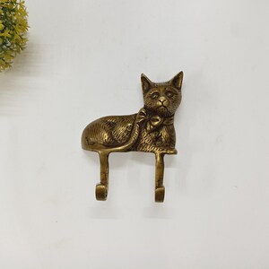 Puede incluir: Un gancho de pared con forma de gato de latón con dos ganchos. El gato está representado en posición sentada con una pajarita. El gancho es un artículo decorativo, adecuado para colgar objetos. El latón tiene un color dorado cálido.