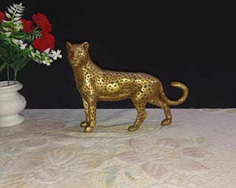 May include: A gold-colored metal sculpture of a leopard. The sculpture is standing on a white surface with a floral pattern.