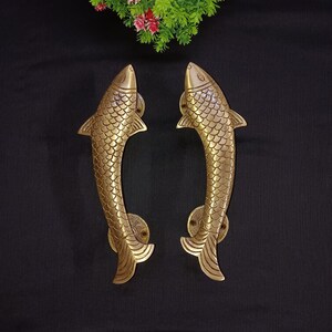 May include: A pair of gold-toned fish-shaped door handles. The handles are detailed with scales and have a curved shape.