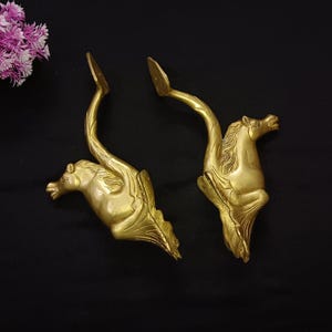 May include: Two gold-toned metal decorative horse head door handles. The handles are in the shape of a horse's head with a curved neck and tail.