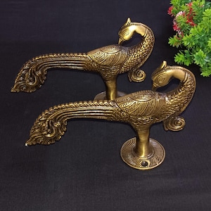 May include: Two antique brass peacock door handles. The handles are detailed with engraved feathers and a curved neck. The handles are mounted on a round base. The background is a dark surface.