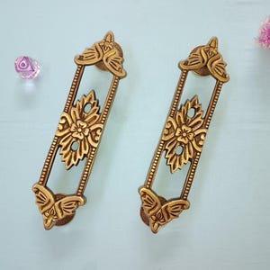 Puede incluir: Dos tiradores de cajón de color dorado con diseños florales ornamentados. Los tiradores tienen una forma rectangular con un diseño de mariposa en la parte superior e inferior.