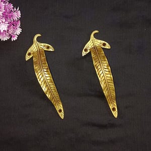 Może przedstawiać: Dwa złote uchwyty do szuflad w kształcie liści. Każdy uchwyt ma długi, smukły korpus ze szczegółowym żyłkowaniem i zakrzywioną górą z dwoma małymi otworami. Uchwyty są umieszczone na ciemnym tle, z klastrem fioletowych kwiatów w lewym górnym rogu.