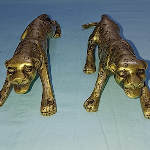 Könnte beinhalten: Zwei goldfarbene Metallstatuen von Leoparden. Die Leoparden befinden sich in einer hockenden Position mit nach vorne gerichteten Köpfen. Die Statuen sind mit Flecken verziert und haben ein realistisches Aussehen.