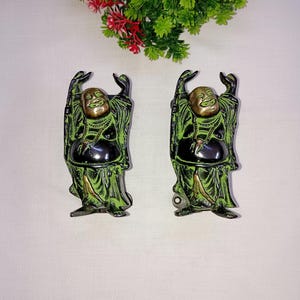 Op de afbeelding: Twee groene en zwarte lachende Boeddha-beeldjes met opgeheven armen. De beeldjes zijn gemaakt van metaal en hebben een gouden rand.
