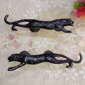 Könnte beinhalten: Zwei schwarze Metall-Türgriffe in Form von Leoparden. Die Griffe sind mit Flecken verziert und haben ein elegantes, modernes Design.