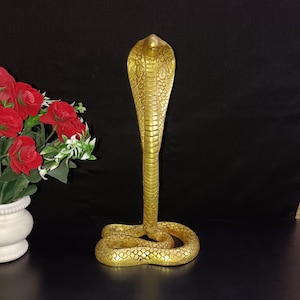 Op de afbeelding: Een gouden metalen beeld van een cobra met zijn kop omhoog en zijn lichaam opgerold. Het beeld is gedetailleerd met schubben en heeft een gladde, gepolijste afwerking.
