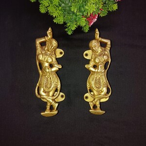 Op de afbeelding: Een paar goudkleurige metalen deurgrepen in de vorm van dansende figuren. De figuren zijn gedetailleerd met ingewikkelde houtsnijwerk en zijn in een traditionele Indiase stijl.