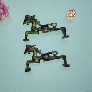 May include: Two ornate black and green metal door handles shaped like horses with intricate details. The handles are on a light blue background.