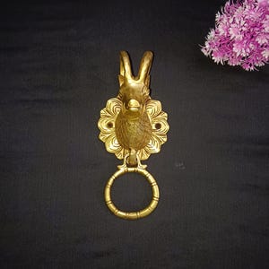 May include: A gold-toned door knocker with a detailed design of a ram's head. The knocker has a ring attached to the bottom for pulling.
