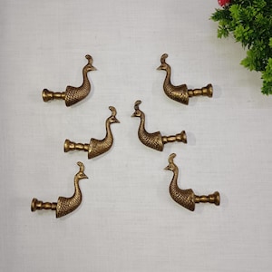 Puede incluir: Seis ganchos decorativos con forma de pavo real de color bronce, dispuestos sobre una superficie blanca. Cada gancho presenta un diseño detallado de pavo real. Los ganchos son probablemente para colgar objetos.