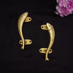 May include: Two gold-toned fish-shaped door handles with a detailed design. The handles are mounted on oval-shaped plates.