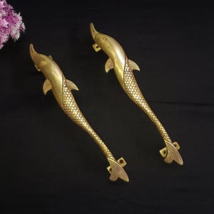 Könnte beinhalten: Zwei goldfarbene Delfin-Türgriffe mit detaillierten Schuppen. Die Griffe sind aus Metall und haben eine polierte Oberfläche.