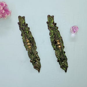 Puede incluir: Dos tiradores de cajón ornamentados de metal verde y dorado con un diseño floral. Los tiradores están orientados verticalmente y tienen un acabado envejecido. También hay un pequeño pomo de cristal transparente con un diseño de remolino púrpura.