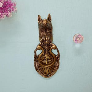 May include: A gold-toned door knocker with a detailed design featuring a lion's head and two peacocks. The knocker is mounted on a pale blue wall.