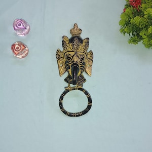 May include: Black metal door knocker with a gold-painted design of a deity's face. The knocker has a circular ring for pulling.