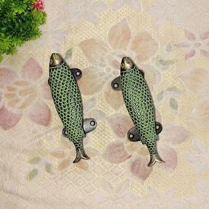 May include: Two green and gold fish-shaped door handles with a textured, scaly design. The handles are made of metal and have a vintage look.
