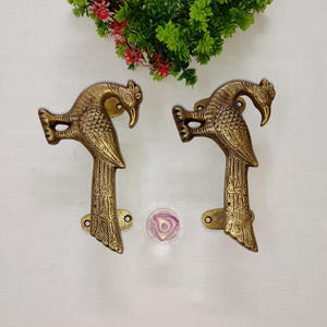 May include: Two brass peacock-shaped door handles. The handles are detailed with feathers and have a golden finish. The handles are mounted on a white surface with a decorative glass orb and greenery in the background.