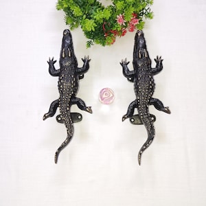 May include: Two dark bronze alligator-shaped door handles. The handles are detailed with textured scales and have a decorative, antique finish. The handles are mounted on a white surface, with a small floral arrangement above.