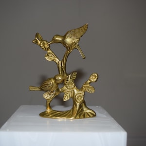 May include: A gold-colored metal sculpture of two birds perched on a tree branch with leaves and flowers. The sculpture is on a white surface and has a detailed design.