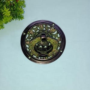 May include: A round, ornate door knocker with a decorative design of flowers and leaves. The knocker is made of brass and has a dark green patina.
