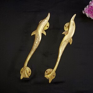 Op de afbeelding: Twee messing deurklinken in de vorm van een dolfijn. De handgrepen zijn goudkleurig, met een gedetailleerd schubbenpatroon op het lichaam en de vinnen. De handgrepen zijn gemonteerd op ronde platen met een textuur. De achtergrond is zwart.