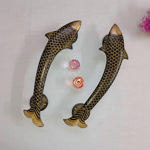 May include: A pair of decorative door handles shaped like fish. The handles are made of brass and have a black and gold finish. The fish have scales and fins.