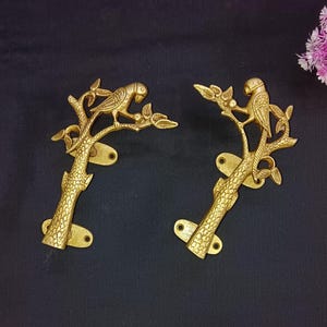 Puede incluir: Dos manijas de puerta de color latón. Cada manija presenta un loro posado en una rama de árbol con hojas. Las manijas tienen un diseño de tronco de árbol texturizado y orificios de montaje. Fondo oscuro.