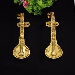 Puede incluir: Un par de tiradores de puerta de color dorado con forma de sitar, un instrumento musical tradicional de la India. Los tiradores están montados sobre un fondo negro.