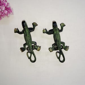 Peut inclure: Deux poignées de porte décoratives en forme de lézard. Les poignées sont en métal et présentent un schéma de couleurs vert et noir avec des accents dorés. Les lézards sont en position de rampement, la queue enroulée.