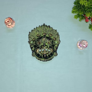 Puede incluir: Una máscara de metal verde y negro con detalles intrincados y una expresión feroz. La máscara tiene un diseño en relieve y parece estar hecha de latón.