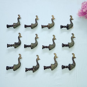 Könnte beinhalten: Zwölf dekorative Wandhaken in Pfau-Form. Jeder Haken besteht aus dunkelbronze-farbenem Metall mit einem goldfarbenen Kopf. Die Haken sind in drei Reihen zu je vier Stück auf weißem Hintergrund angeordnet. Ein Strauß rosa Blumen befindet sich in der oberen rechten Ecke.