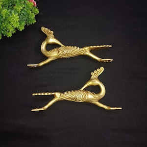 May include: Two brass peacock figurines with intricate detailing, set against a black background. The peacocks are in a running pose, with their tails spread and bodies curved. The figurines have a golden hue and are likely decorative items.