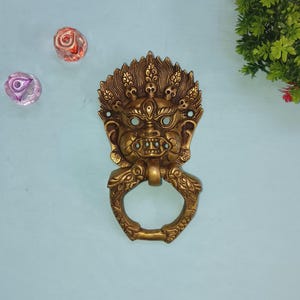 Peut inclure: Un heurtoir de porte en laiton détaillé en forme de visage féroce avec une poignée en anneau. Le heurtoir présente des détails complexes, dont une couronne de crânes. Deux pierres décoratives en verre sont visibles.