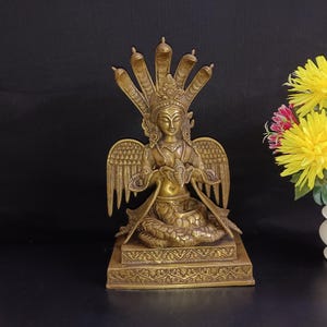 Könnte beinhalten: Eine goldfarbene Messingstatue einer Gottheit mit einem schlangenartigen Kopfschmuck und Flügeln, die auf einem Sockel mit aufwendigen Schnitzereien sitzt. Die Statue hält eine kleine Figur in ihren Händen.