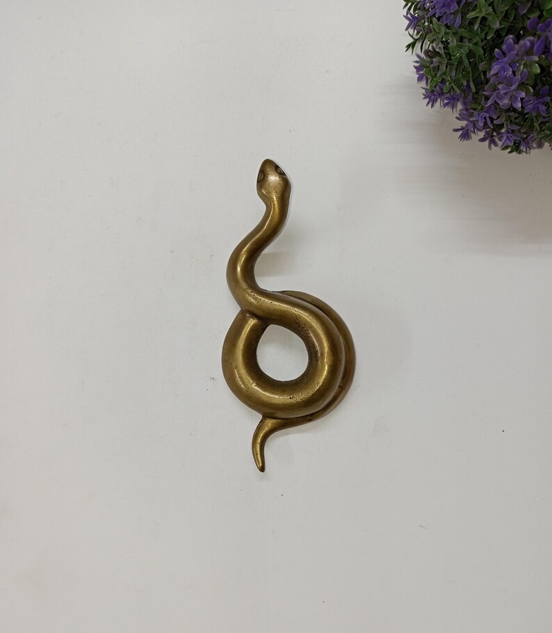 May include: A brass snake ornament with a coiled body and raised head. The sculpture has a smooth, polished finish and is set against a white background. The snake's form is detailed, showcasing its curves and the shape of its head.