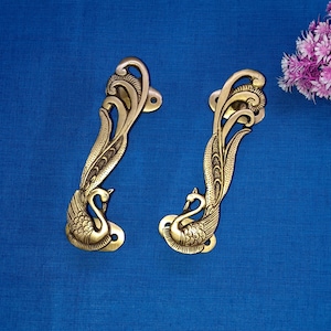 May include: Two ornate brass door handles shaped like peacocks with intricate details. The handles are on a blue background.