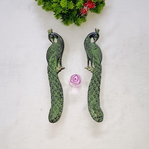 Może przedstawiać: Dwa zielone dekoracyjne uchwyty w kształcie pawia z czarnymi akcentami i teksturowanym wykończeniem. Uchwyty są ustawione pionowo, z małym, przezroczystym, różowym, okrągłym przedmiotem pomiędzy nimi. Uchwyty są prawdopodobnie do szafki lub szuflady.