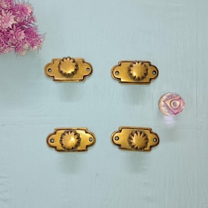May include: Four antique brass drawer pulls with a floral design, arranged on a light blue surface. A decorative glass knob is also present. A cluster of pink flowers is in the upper left corner.