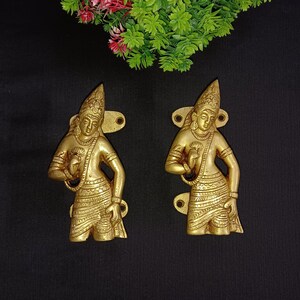 Könnte beinhalten: Ein Paar goldfarbene Türklinken in Form von Figuren in traditioneller indischer Kleidung. Die Griffe sind mit aufwendigen Schnitzereien verziert und haben ein dekoratives Design.
